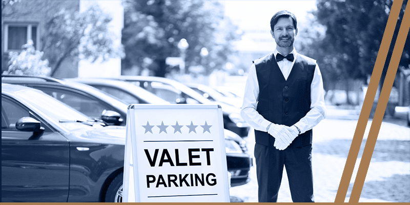 Valet attendant in uniform stands next to a "Valet Parking" sign in a parking lot with parked cars and trees in the background.